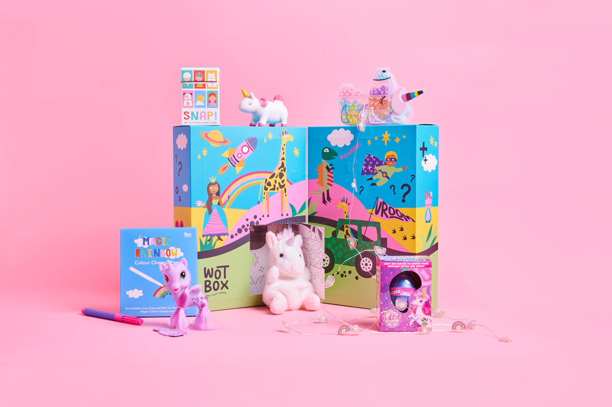 A colorful children's toy set arranged on a pink studio background, including two illustrated boxes labeled 'WOTBOX', a small unicorn plush, a pink unicorn figurine, a small book or card, and a pink accessory. No people, animals, or real environment are present, so this is GIFT BOX product.