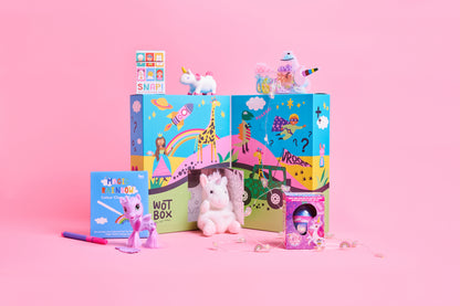 A colorful children's toy set arranged on a pink studio background, including two illustrated boxes labeled 'WOTBOX', a small unicorn plush, a pink unicorn figurine, a small book or card, and a pink accessory. No people, animals, or real environment are present, so this is GIFT BOX product.