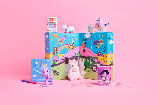 A colorful children's toy set arranged on a pink studio background, including two illustrated boxes labeled 'WOTBOX', a small unicorn plush, a pink unicorn figurine, a small book or card, and a pink accessory. No people, animals, or real environment are present, so this is GIFT BOX product.